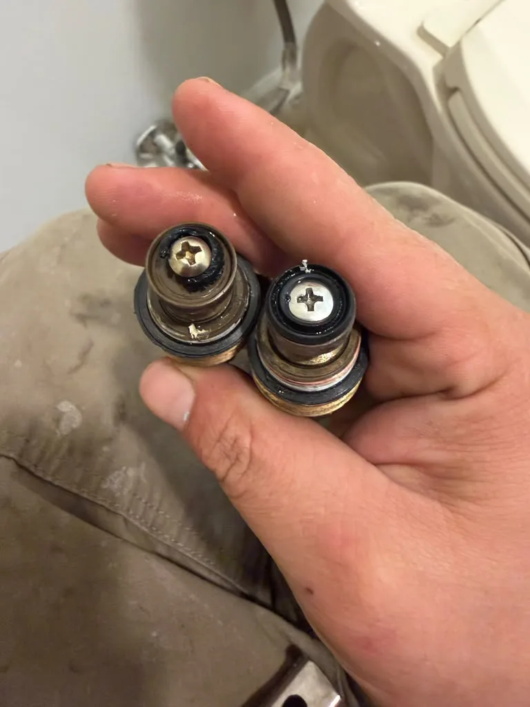 A hand holding two removed faucet valve cartridges next to a toilet