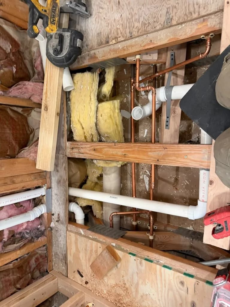 Open wall cavity showing new copper and PVC pipe rough-in with insulation and tools nearby
