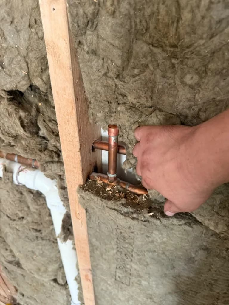 A hand positioning a copper pipe stub-out inside a wall between fiberglass insulation