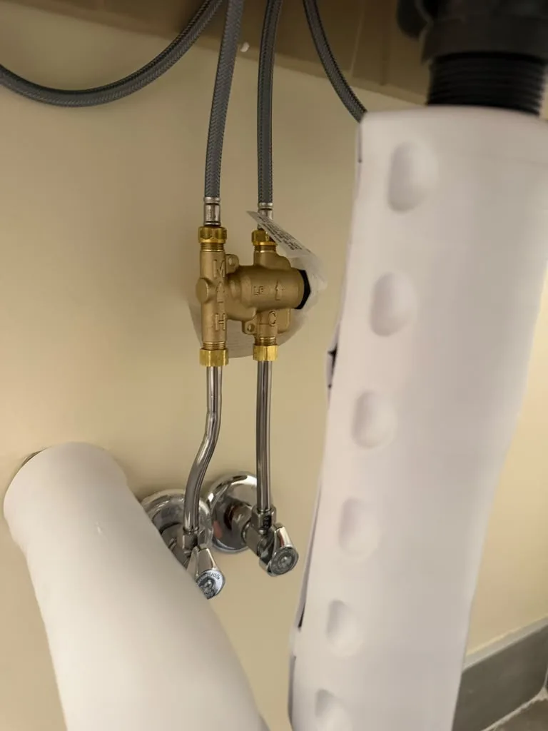 Close-up of a brass hot and cold water mixing (tempering) valve installed under a sink with braided supply lines