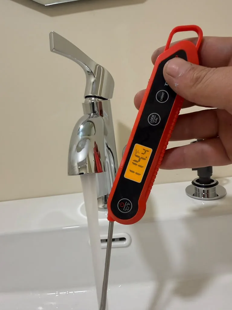 Digital thermometer measuring the temperature of hot water running from a bathroom faucet, reading in the low 100s Fahrenheit